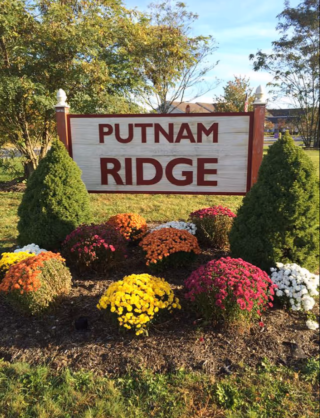 A wooden sign with red letters reading 'PUTNAM RIDGE' surrounded by colorful flowers and two green shrubs on either side, set outdoors with trees and a building visible in the background under a clear sky.