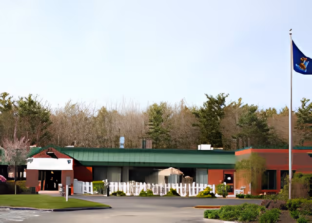 Exterior view of a single-story building with a green roof and red walls, surrounded by trees and bushes. A flagpole with a flag is visible on the right side, and there is a paved driveway leading to the entrance.