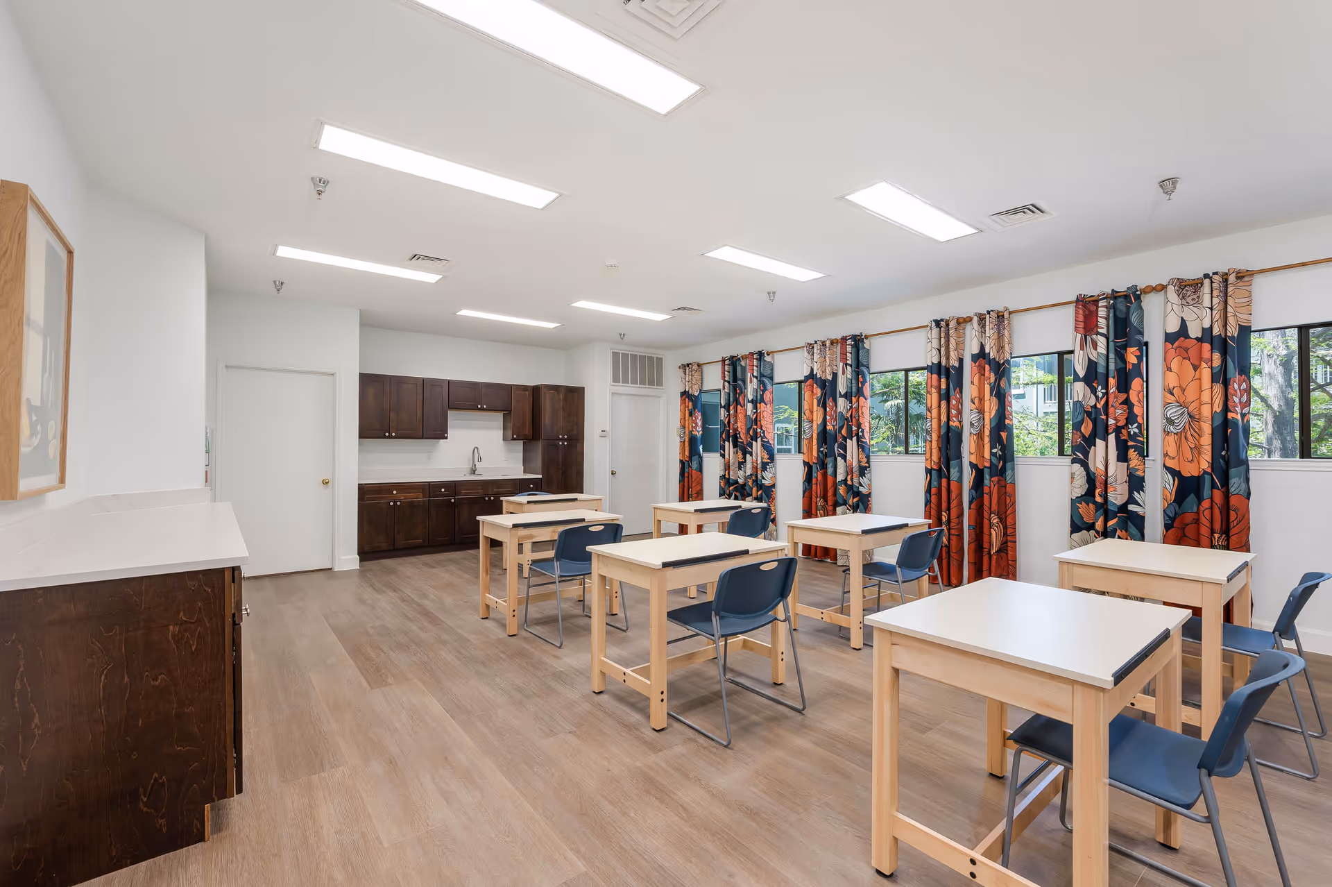 A bright room with several small wooden tables and blue chairs arranged in rows. The room has large windows with colorful floral curtains, wooden flooring, and a kitchenette with dark wood cabinets and a sink at the back.
