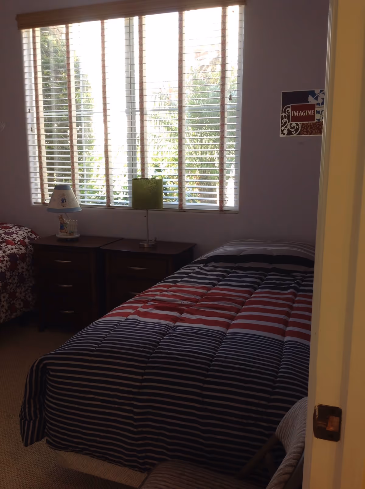 Small bedroom with a striped twin bed, two nightstands with lamps, a large window with blinds and wall decor.
