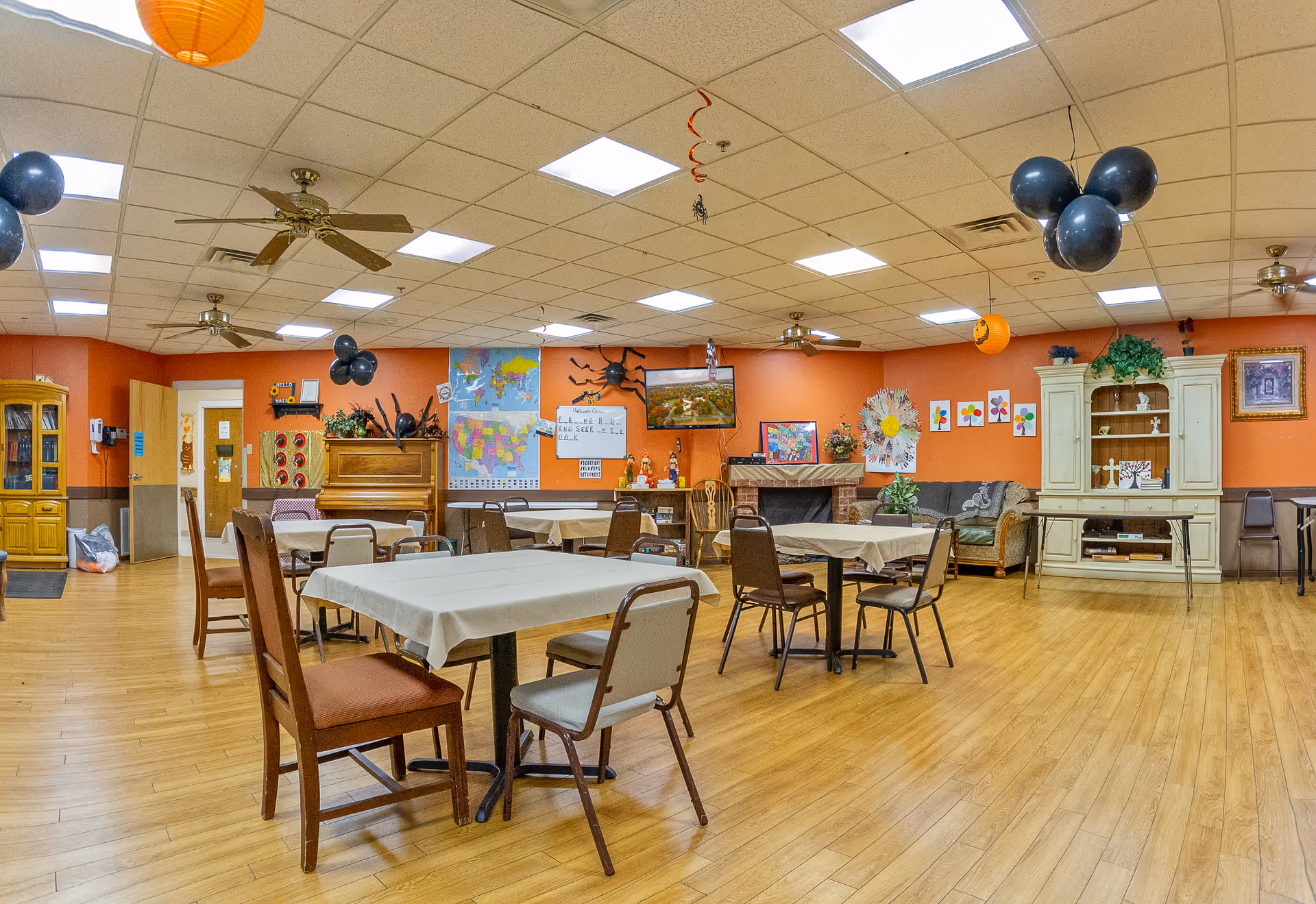 A spacious common room with several tables and chairs arranged for group seating. The walls are painted orange and decorated with maps, artwork, and Halloween-themed decorations including black balloons and hanging orange lanterns. There is a piano against one wall, a TV mounted above a fireplace, and a white cabinet with shelves displaying various items. The floor is wooden, and ceiling fans and fluorescent lights are visible on the ceiling.