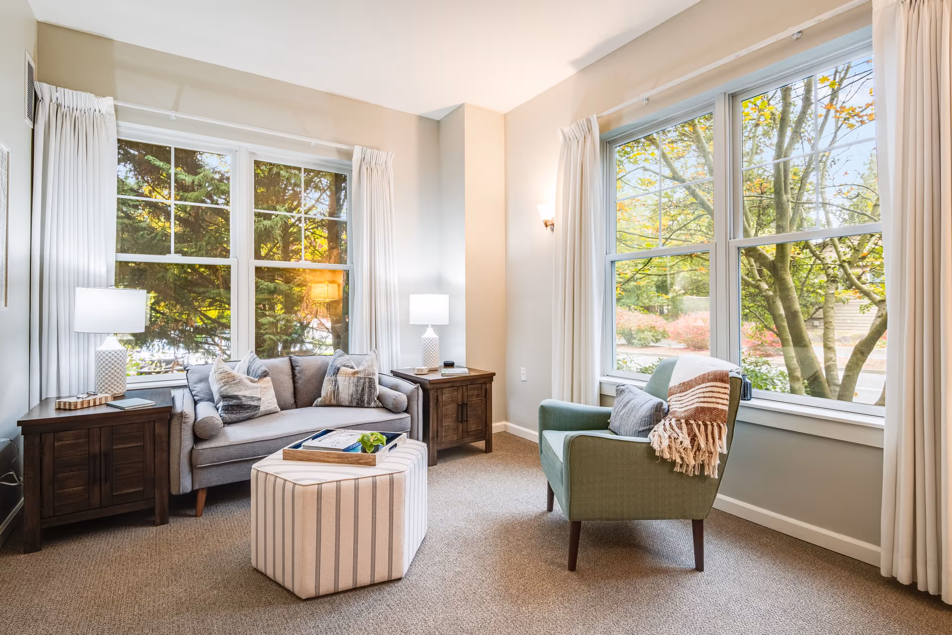 Bright, well-lit living room with a sofa, armchair, ottoman, side tables and large windows overlooking trees.