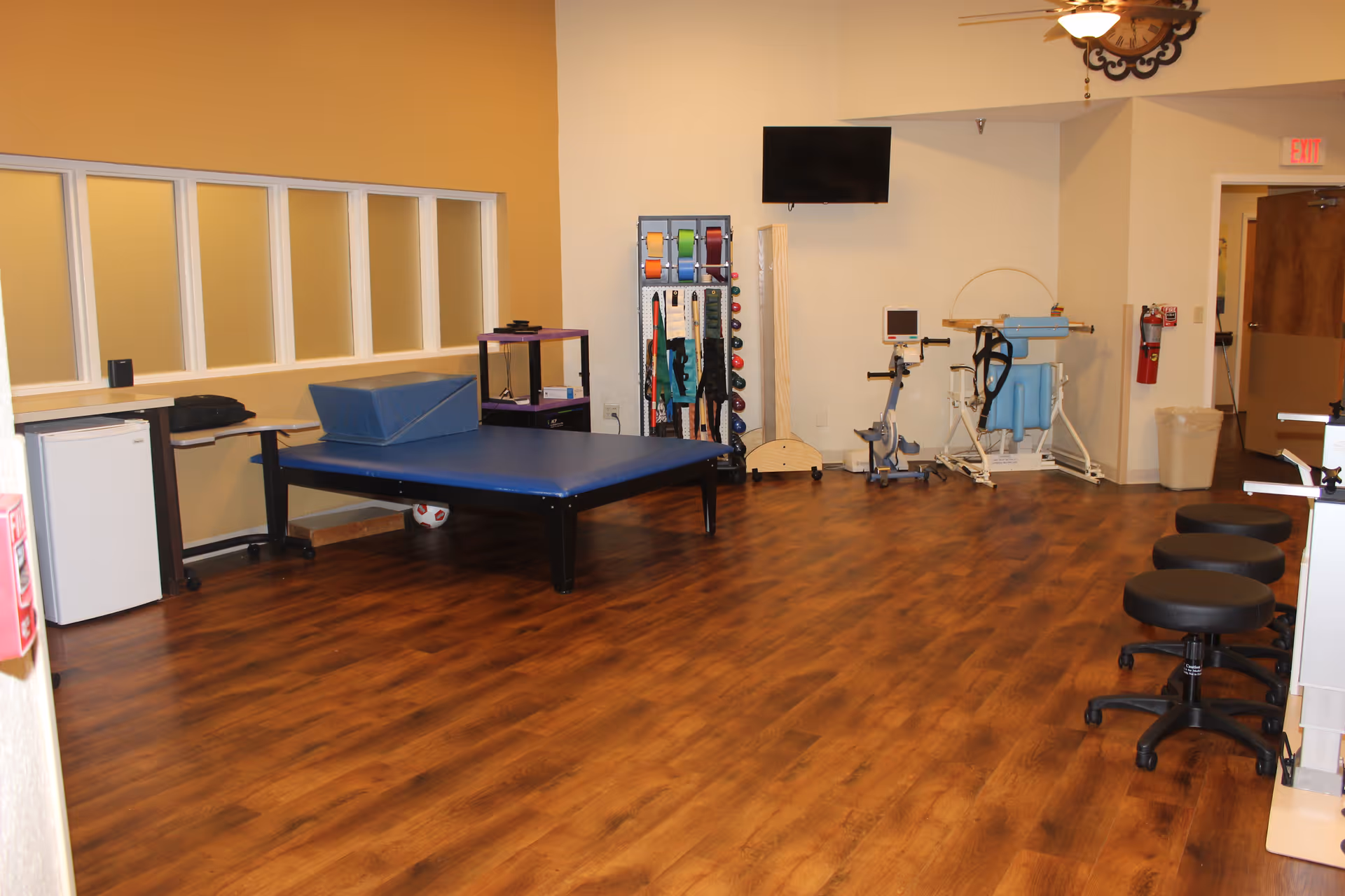 A rehabilitation therapy room with wooden flooring, a blue therapy table with a wedge cushion, exercise equipment including resistance bands and weights on a rack, a stationary bike, stools on wheels, a wall-mounted TV, and a wall clock. The room has beige and white walls with a door and an exit sign in the background.