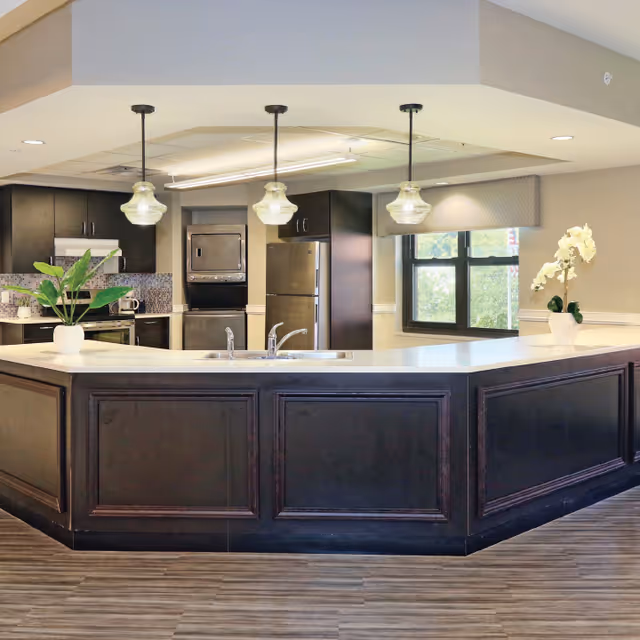 Modern kitchen area with a large dark wood island countertop featuring a sink and faucet. Three pendant lights hang above the island. In the background, there are dark wood cabinets, a stainless steel refrigerator, a microwave, and a window with a beige valance. Two potted plants are placed on the countertop, one with green leaves and the other with white flowers.