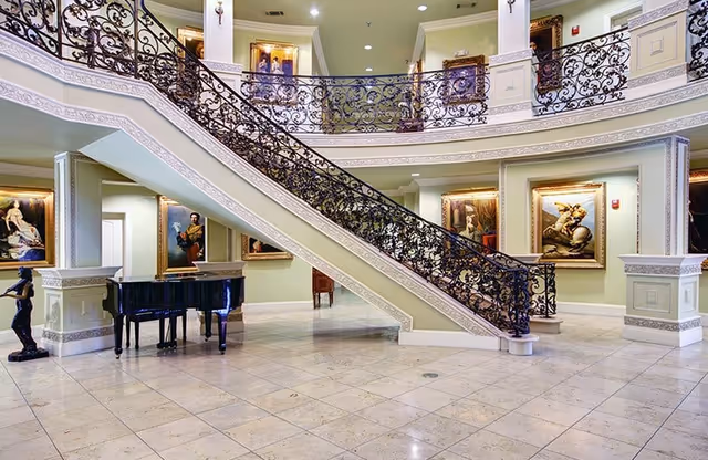 Grand two-story lobby with an ornate wrought-iron staircase, marble floors, framed paintings, and a black grand piano.