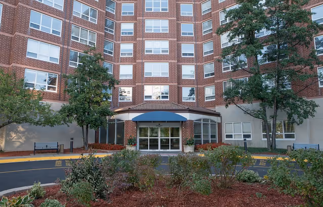 Front exterior view of a multi-story brick building with large windows and a blue awning over the main entrance. There are trees, shrubs, and landscaped areas in front of the building along with benches on either side of the entrance.