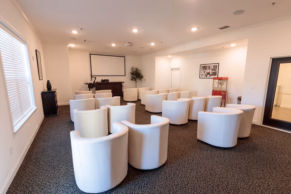 A small theater-style room with white swivel chairs arranged in rows facing a projector screen. The room has white walls, a carpeted floor, a popcorn machine in the corner, a framed black and white photo on the wall, and a window with blinds on the left side.