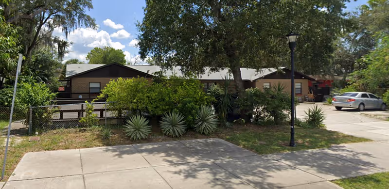 Exterior view of a single-story building partially obscured by trees and bushes, with a sidewalk and a parked silver car visible on the right side under a partly cloudy sky.