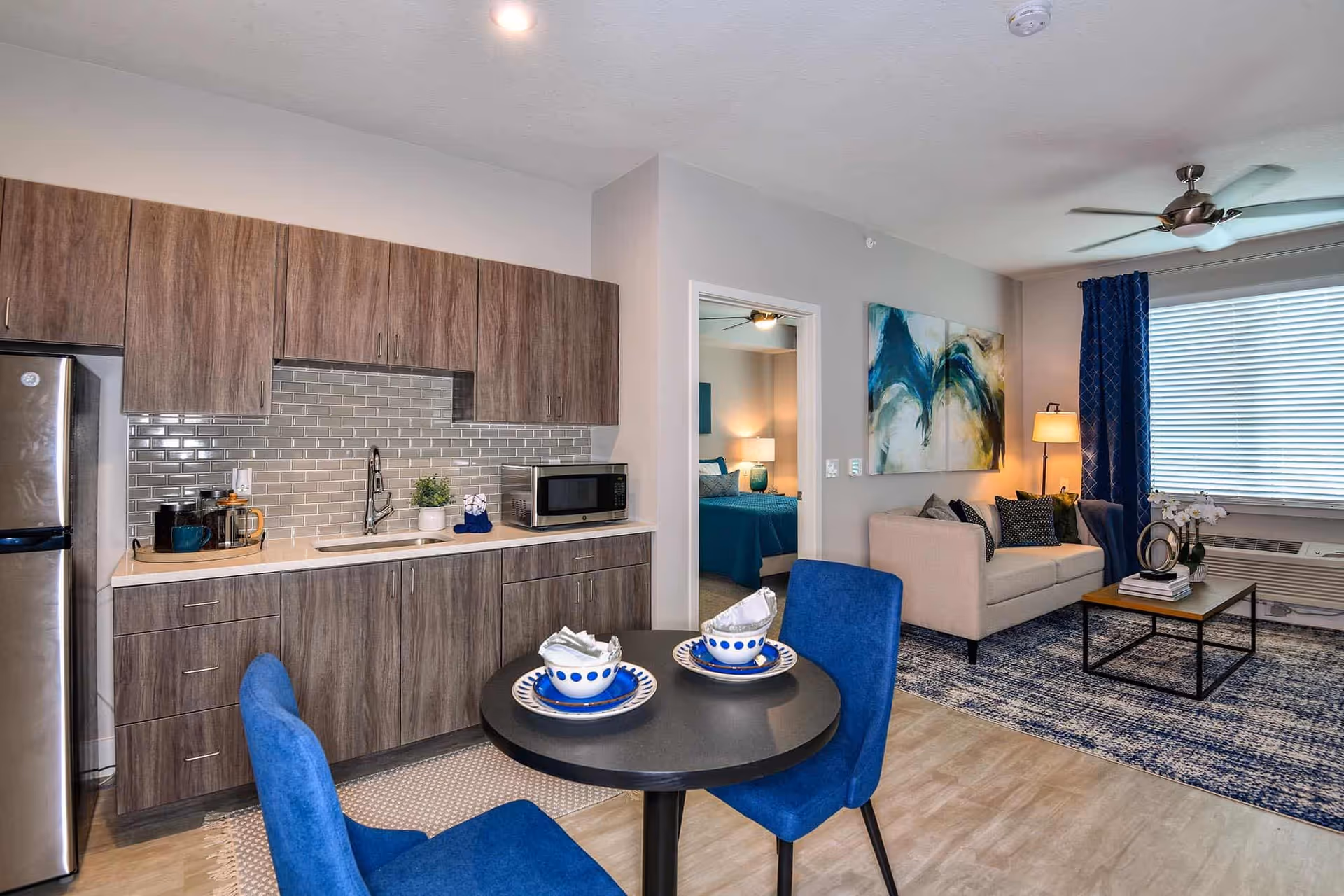 Bright open-plan apartment with a kitchenette, small round dining table with blue chairs, and a living area with a sofa and artwork.