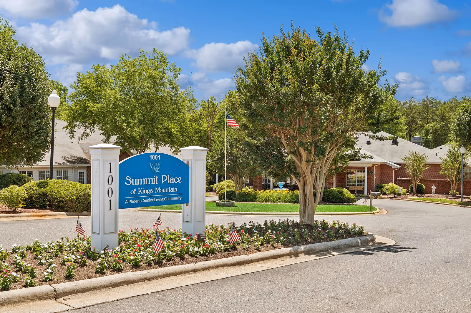 Entrance sign for Summit Place of Kings Mountain, a senior living community, surrounded by a landscaped flower bed with small American flags, trees, and a brick building in the background under a partly cloudy sky.