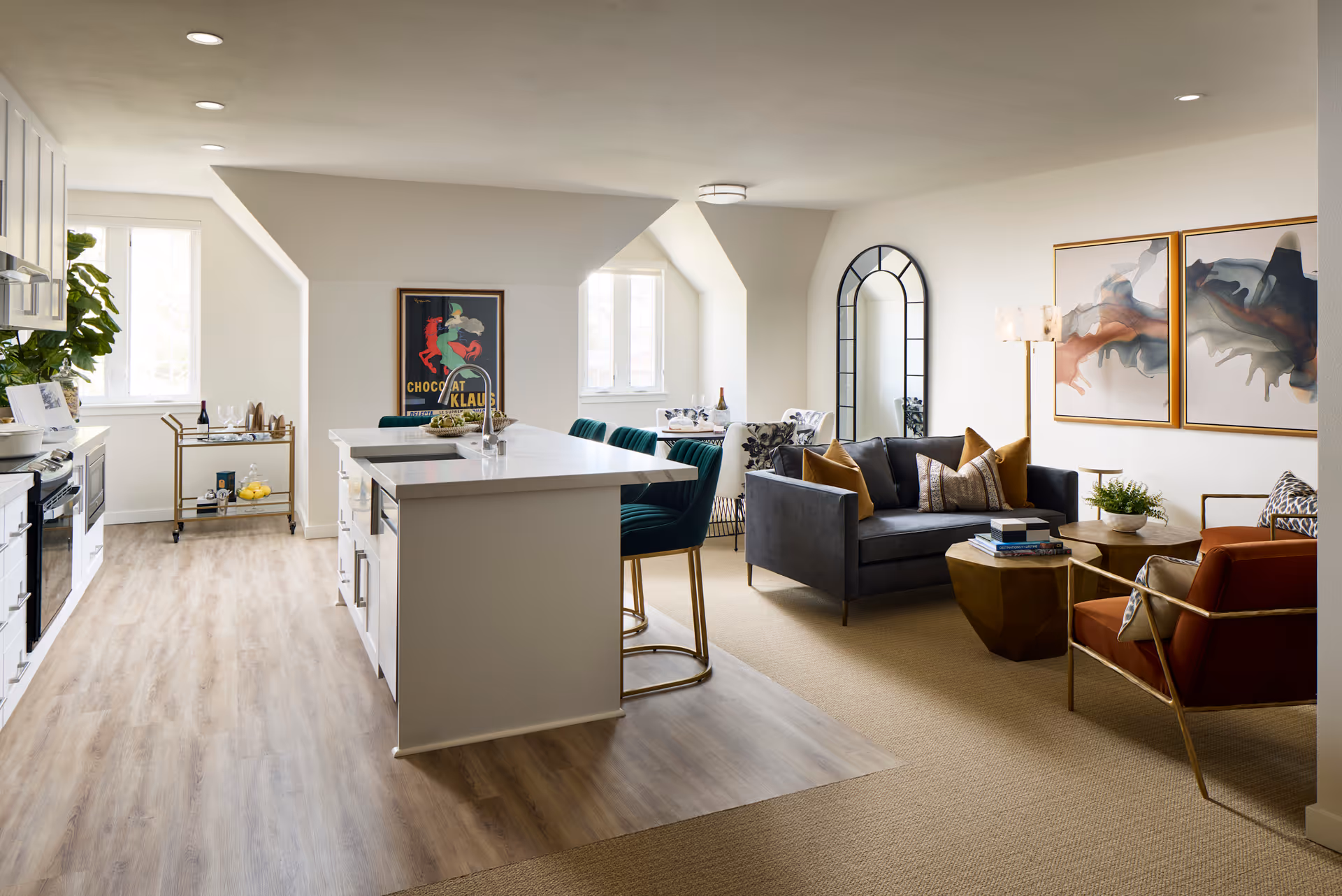 A modern open-concept living space featuring a kitchen with white cabinetry and a large island with green bar stools, adjacent to a cozy living room with a dark gray sofa, orange armchair, gold-toned coffee tables, and abstract wall art. The space is well-lit with natural light from windows and has light wood flooring in the kitchen area transitioning to carpet in the living area.