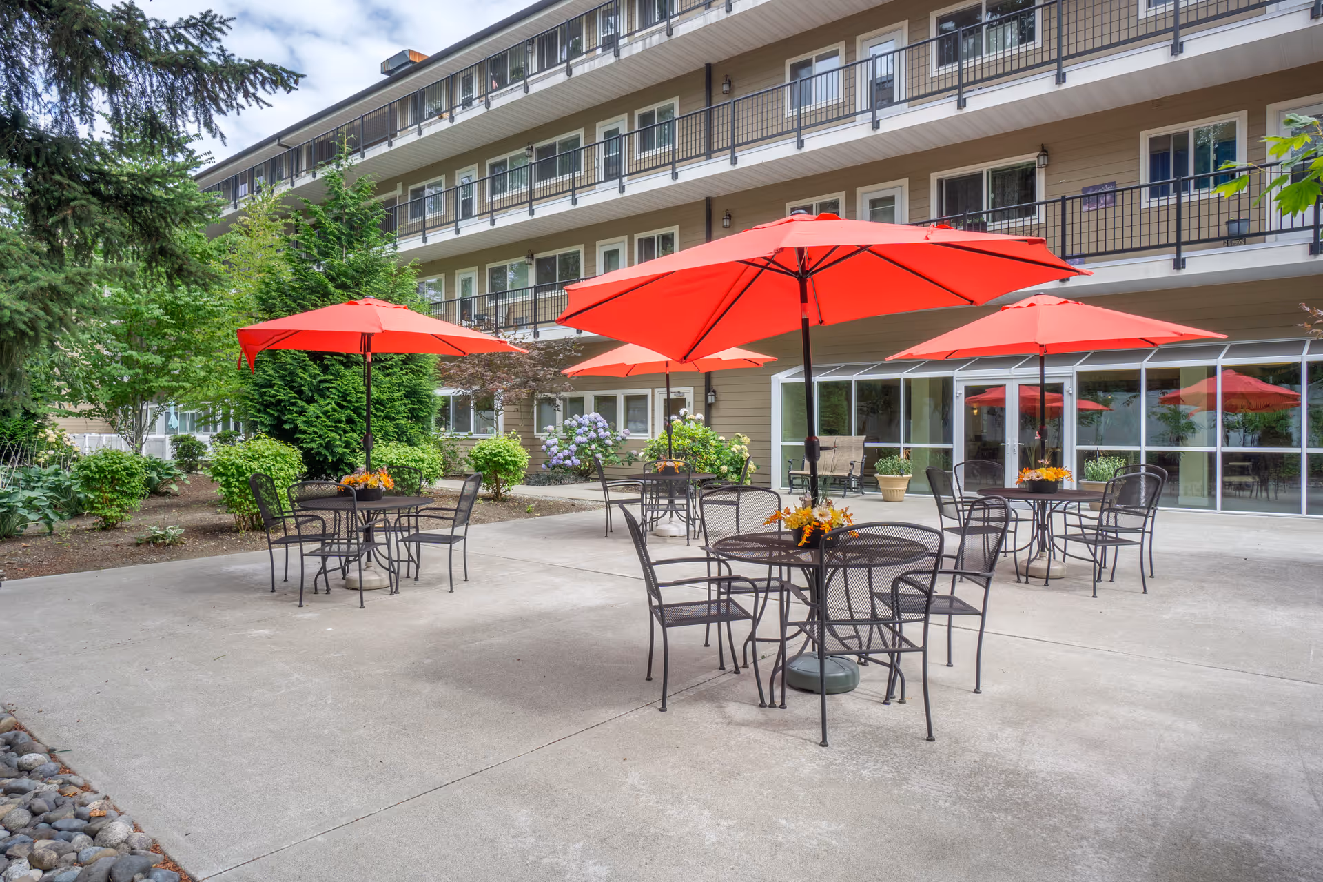 Outdoor patio area at Brookdale Allenmore IL with several black metal tables and chairs, each shaded by large red umbrellas. The patio is surrounded by greenery and flowers, with a multi-story building in the background featuring balconies and large windows.