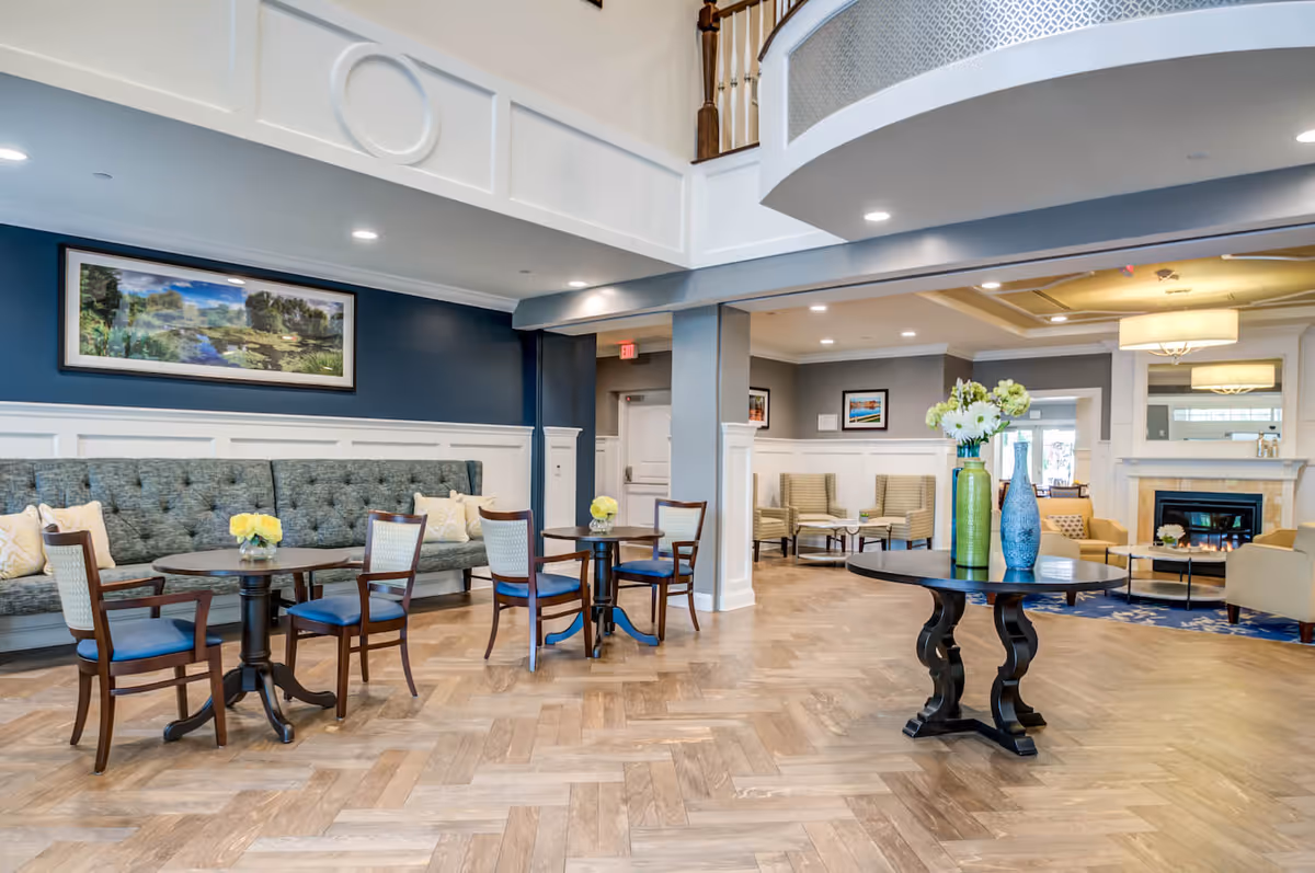 A spacious and well-lit senior living facility common area with wooden herringbone flooring, multiple seating arrangements including a long tufted bench with cushions, wooden chairs with blue seats, and beige armchairs around a fireplace. The walls are painted in blue and gray tones with white wainscoting, and there are framed landscape paintings on the walls. A round table in the center holds decorative vases with flowers. The ceiling has recessed lighting and a balcony railing above.