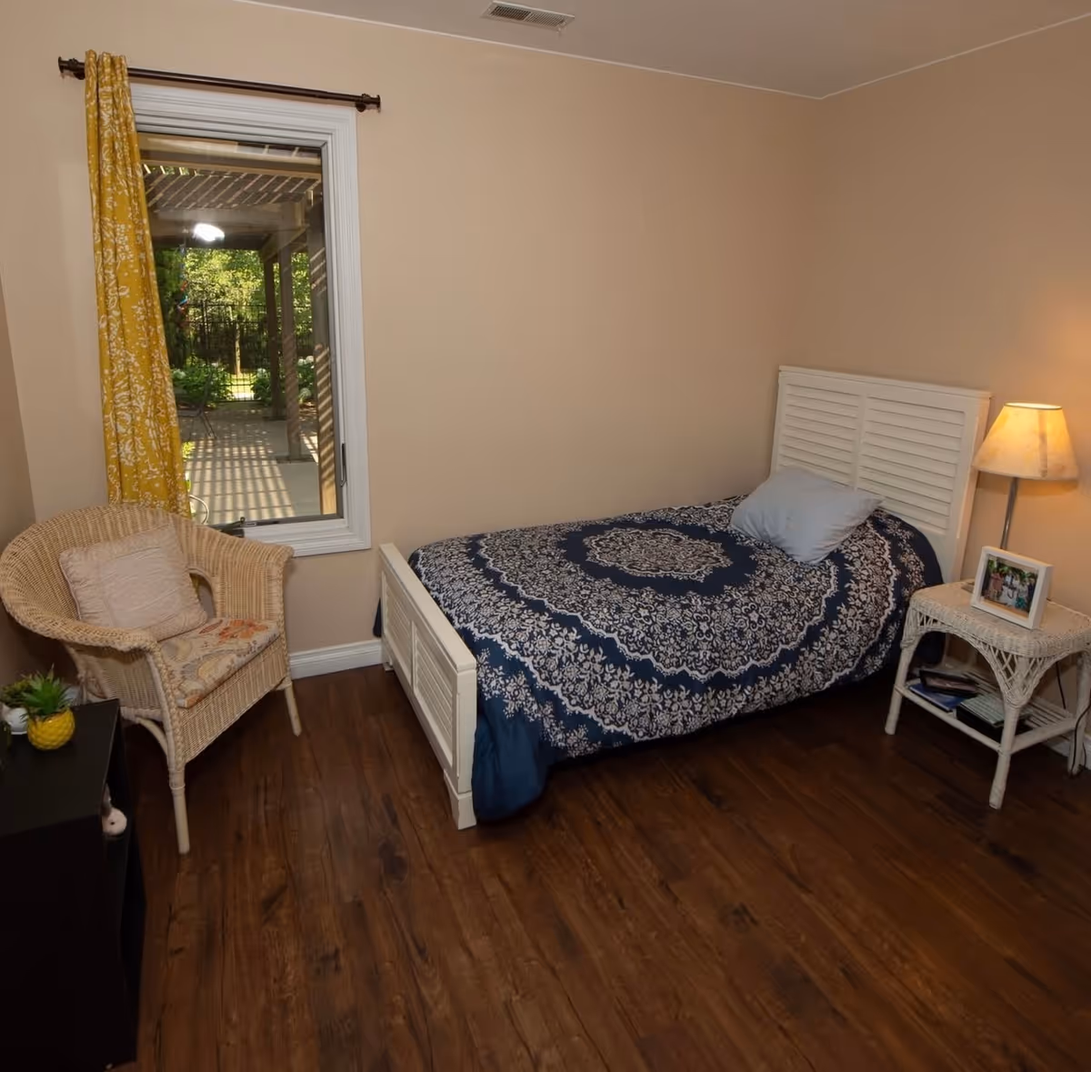 Small bedroom with a single bed covered in a blue patterned quilt, a wicker chair by a window, and a bedside table with a lamp.