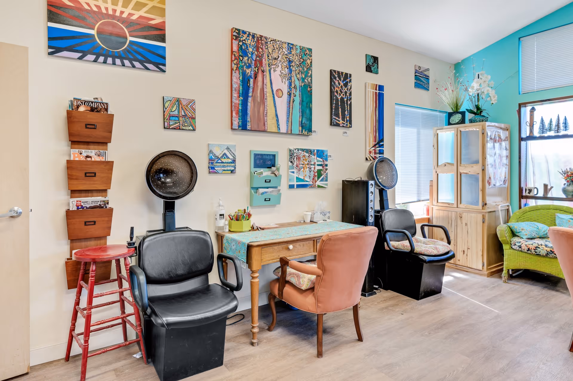 Bright interior common room with salon hair-drying chairs, a wooden table, colorful wall art, and cozy seating by the windows.