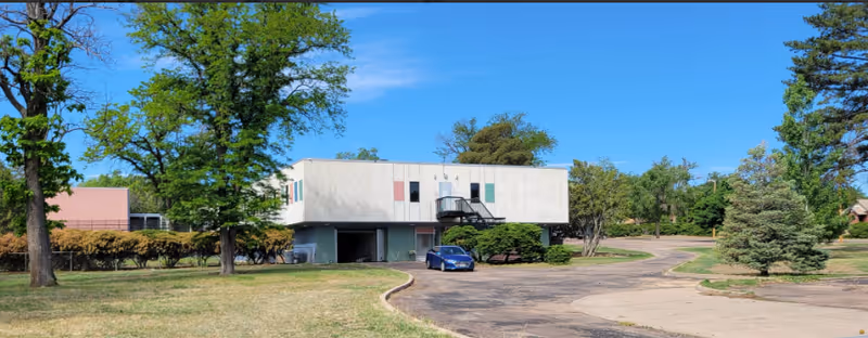 Two-story rectangular building with a covered parking area, a blue car in the driveway, and trees under a clear blue sky.