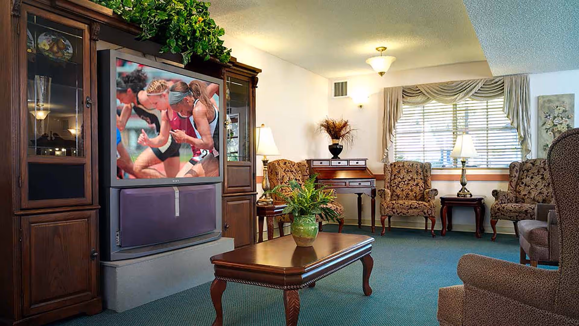 A cozy living room with a large wooden entertainment center housing a television showing a sports event. The room features patterned armchairs, a wooden coffee table with a green plant, a side table with a lamp, a window with drapes, and a small wooden desk with a vase of dried flowers.
