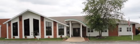 Single-story assisted living building with brick and white siding, tall triangular windows and a central arched entrance, front lawn and a tree.