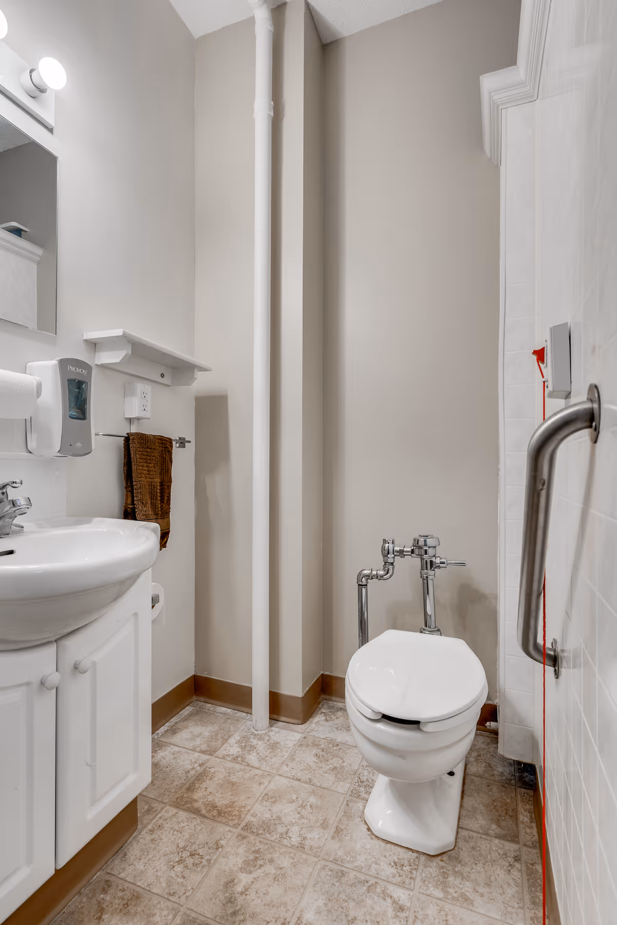 Small accessible bathroom with a toilet, sink and vanity, grab bar, soap dispenser, and tiled floor.