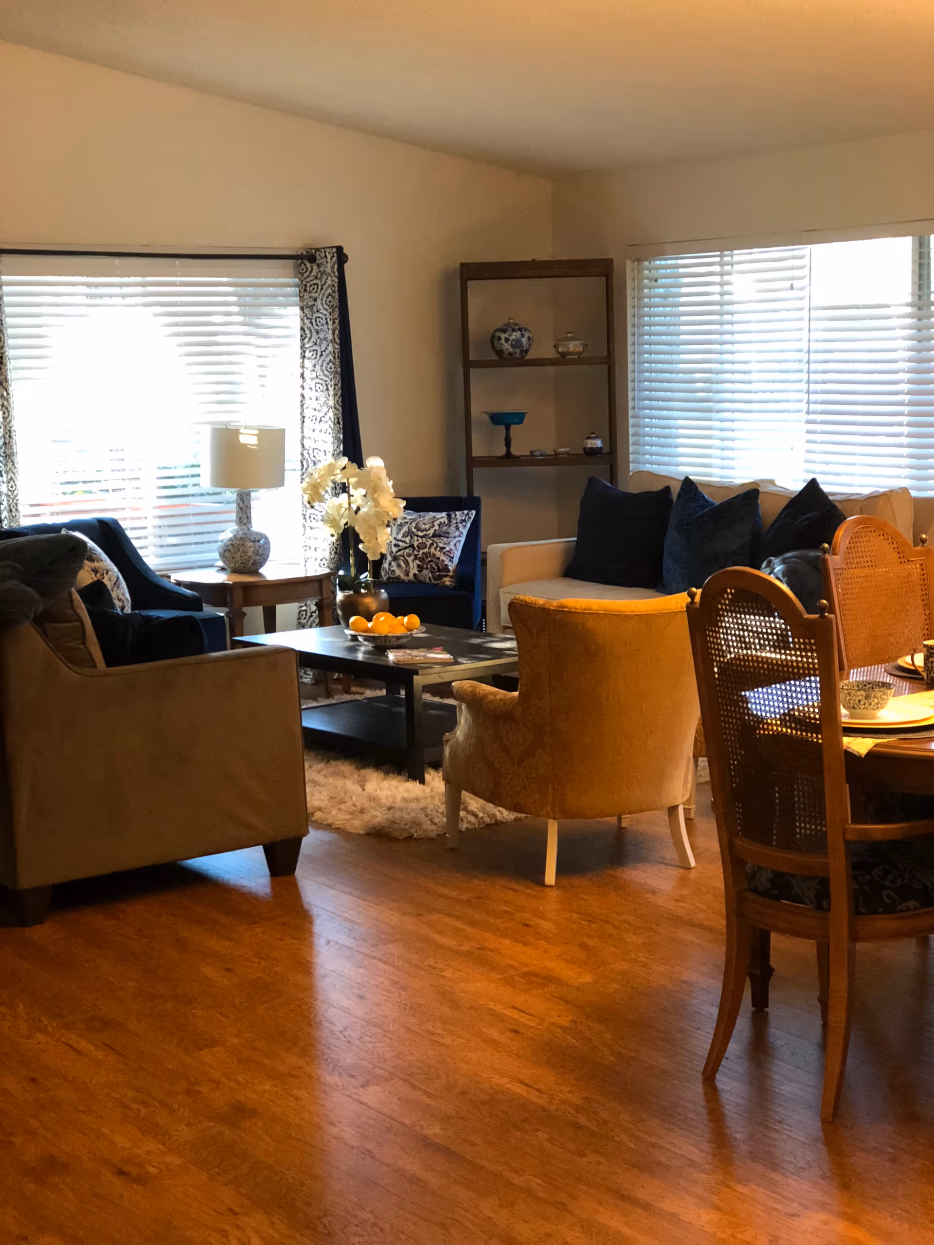 A cozy living room with wooden flooring, featuring a beige armchair, a yellow armchair, a white sofa with dark cushions, and a blue armchair near a window with white blinds and patterned curtains. A coffee table with a bowl of oranges and a vase with white flowers sits in the center. A wooden dining table with chairs is partially visible on the right side, and a wooden shelf with decorative items is against the wall.
