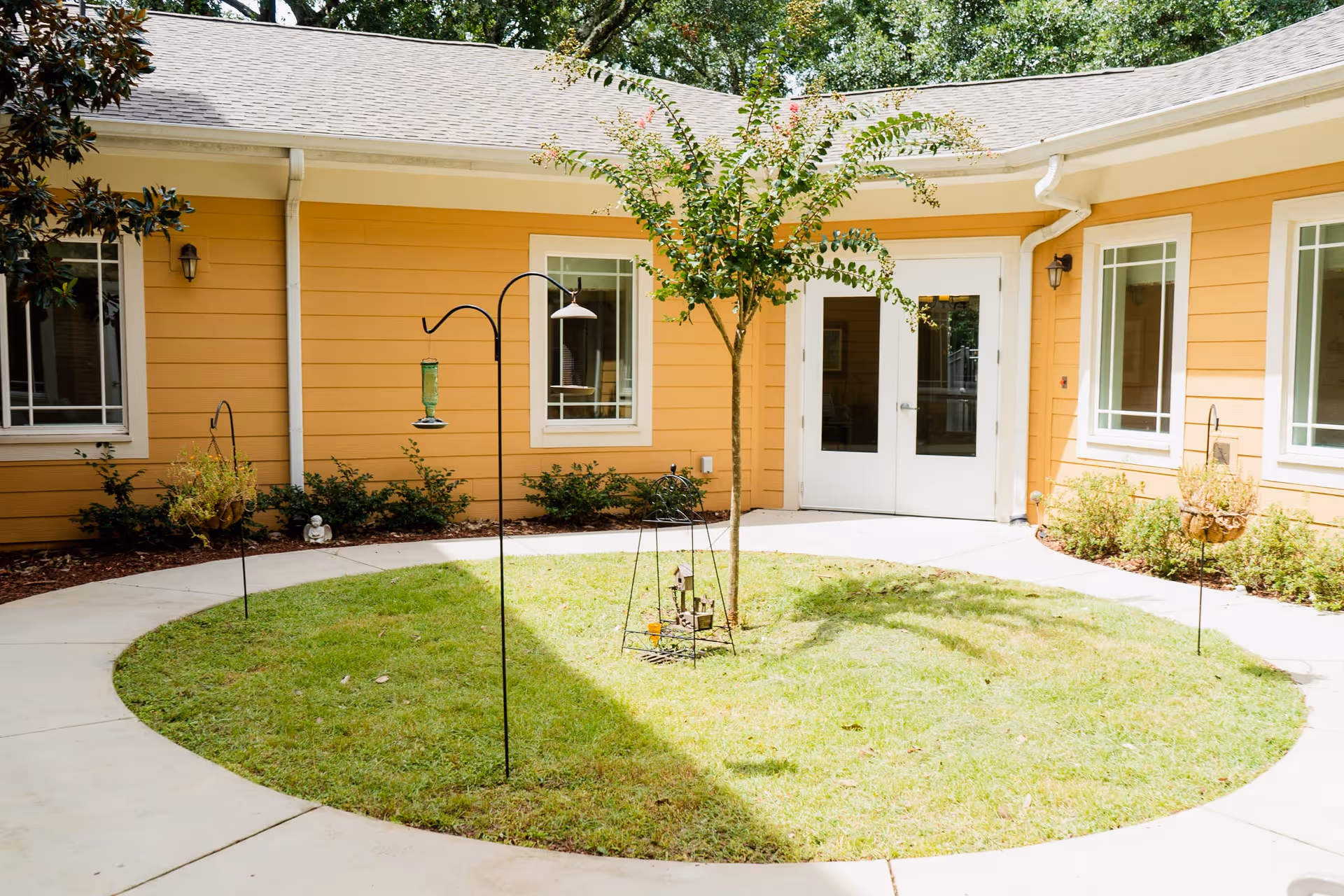 A circular courtyard with a small tree in the center surrounded by a grassy area and a concrete walkway. The courtyard is enclosed by a yellow building with white trim, featuring several windows and a double door entrance. There are bird feeders and small garden decorations around the tree.