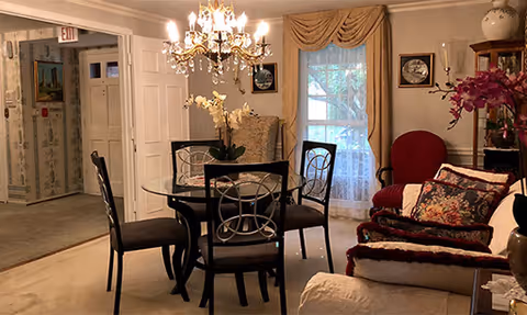 Cozy assisted-living dining area with a round glass table and chairs, chandelier, and an adjacent sitting area with sofa and armchairs.