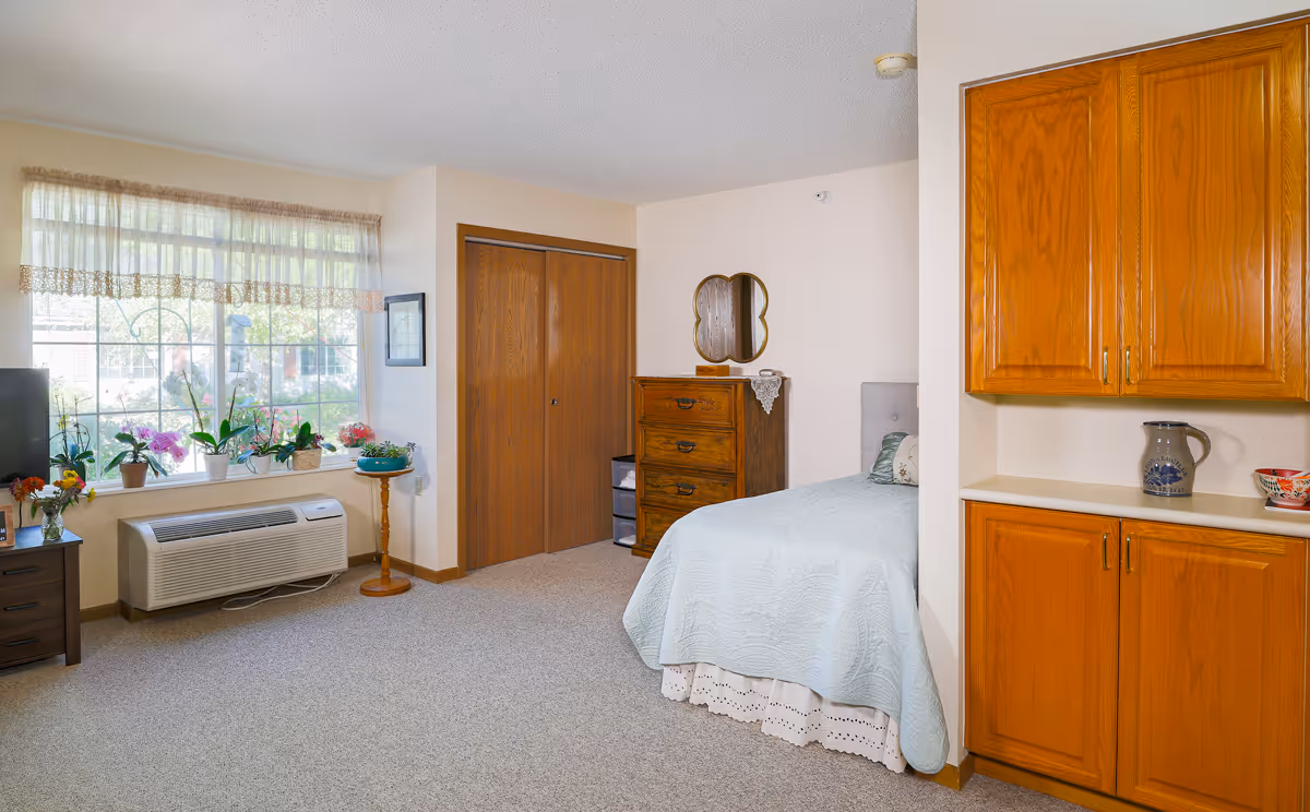 A cozy bedroom in an assisted living facility featuring a bed with a light blue quilt, a wooden dresser with a decorative mirror above it, a large window with sheer curtains and several potted plants on the windowsill, a TV on a small stand, and wooden cabinets on the right side.