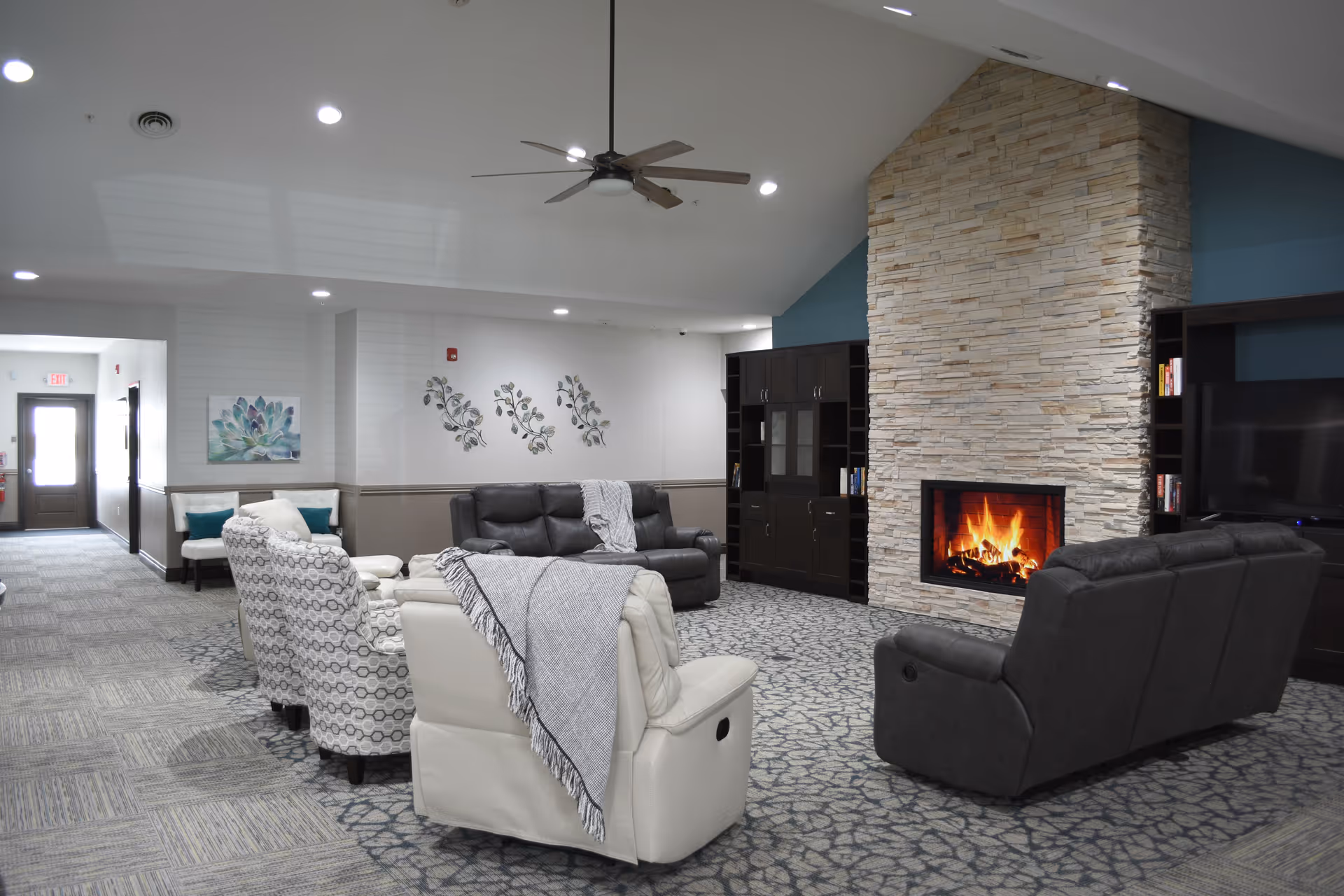 A spacious living room area with a stone fireplace featuring a burning fire. The room has a high vaulted ceiling with recessed lighting and a ceiling fan. There are several comfortable chairs and sofas arranged around the fireplace, including a white recliner with a gray throw blanket, patterned armchairs, and dark leather sofas. The walls are decorated with floral metal art and a painting, and there is a dark wood entertainment center with a TV and bookshelves.