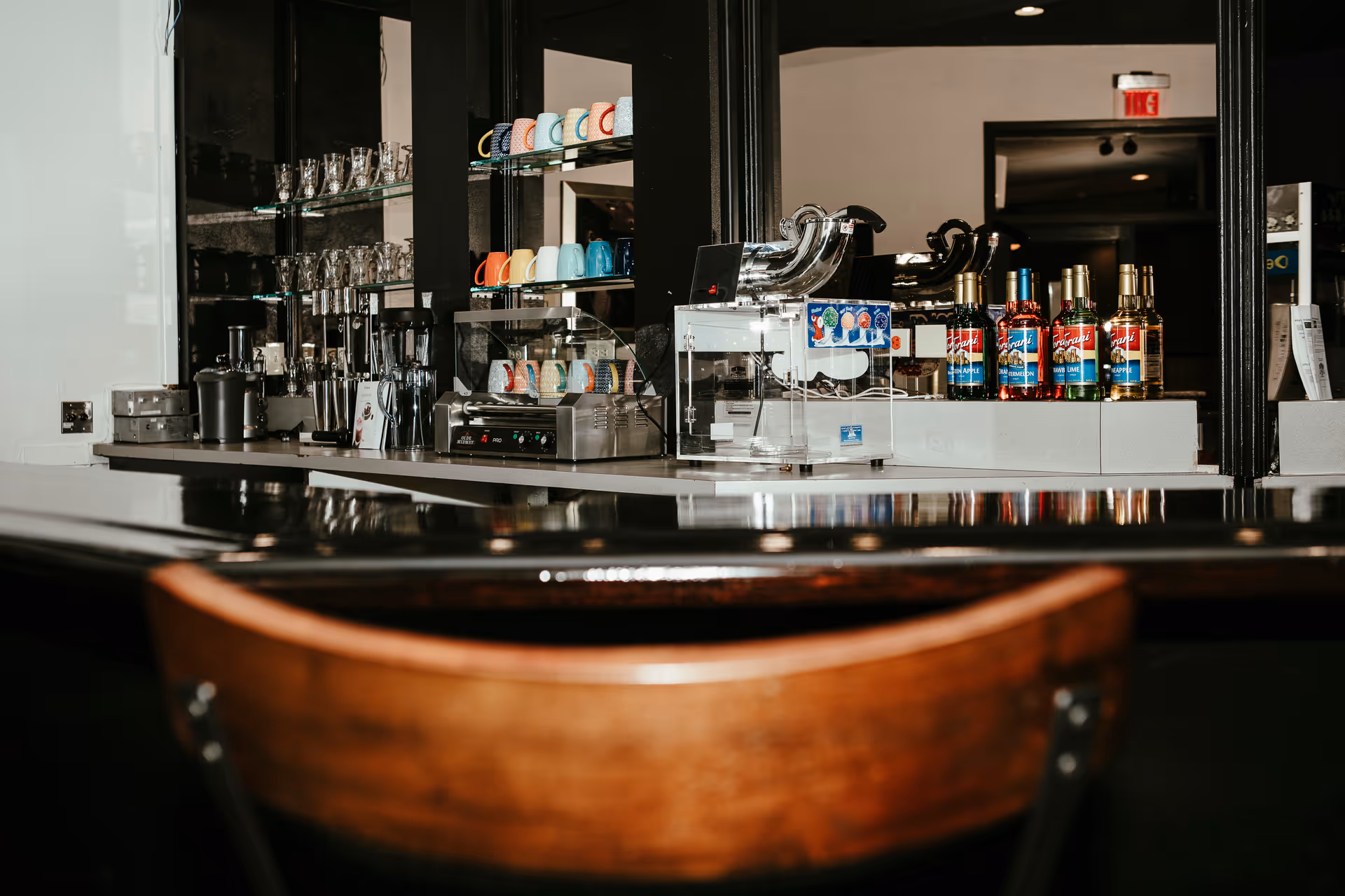 A countertop area with various coffee and beverage supplies including colorful mugs, glassware, a coffee machine, syrup bottles, and a clear container with a dispenser. A wooden chair is partially visible in the foreground.