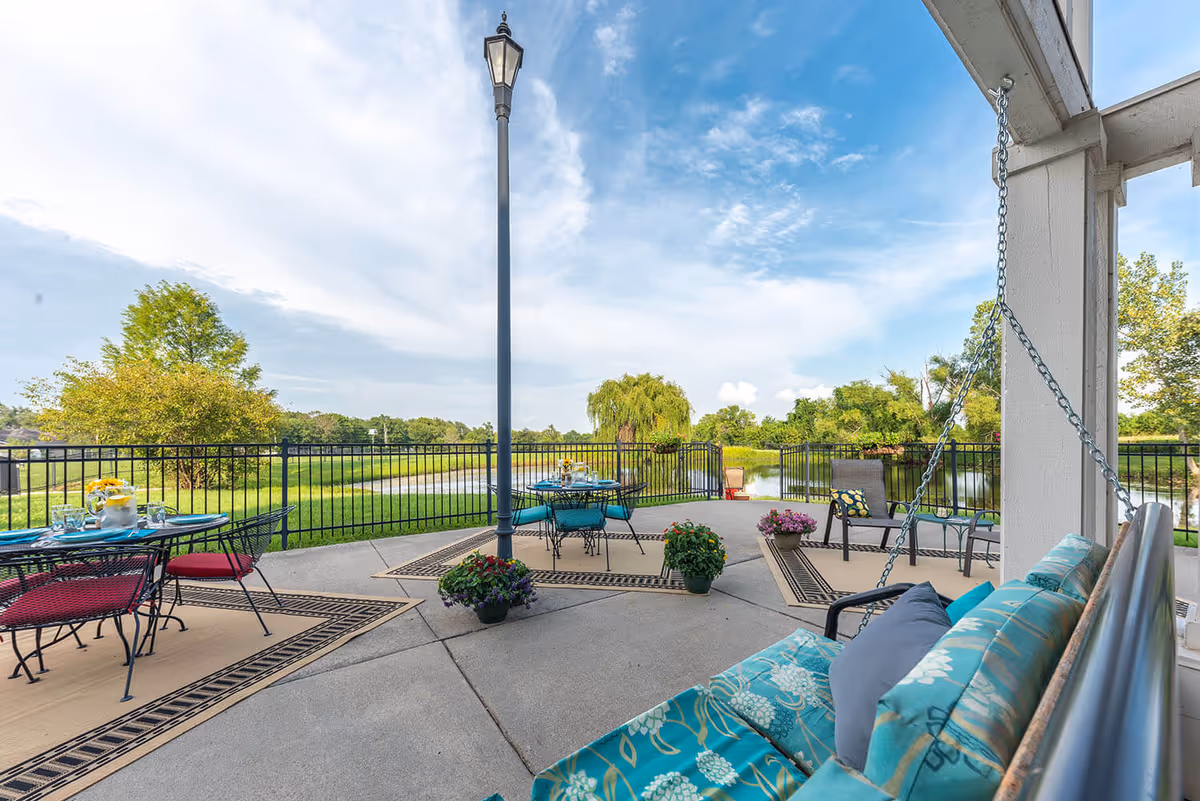 Outdoor patio area with metal tables and chairs, some with red cushions, and a cushioned swing bench with blue patterned pillows. The patio overlooks a grassy area with trees and a pond, under a partly cloudy blue sky.