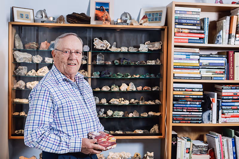 An older man smiling and holding a rock specimen in a room with glass display cases of minerals and a bookshelf.