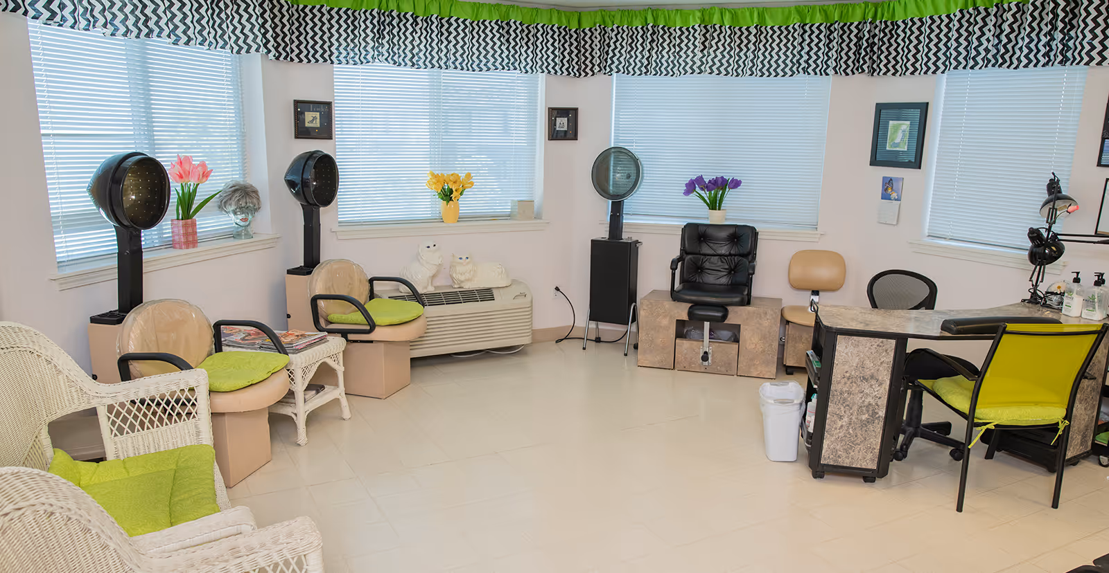 A bright salon area with multiple vintage hair dryers, wicker chairs with green cushions, a reception desk with two chairs, and windows with blinds and patterned valances. There are flowers in vases on the windowsills and some decorative items on the walls.