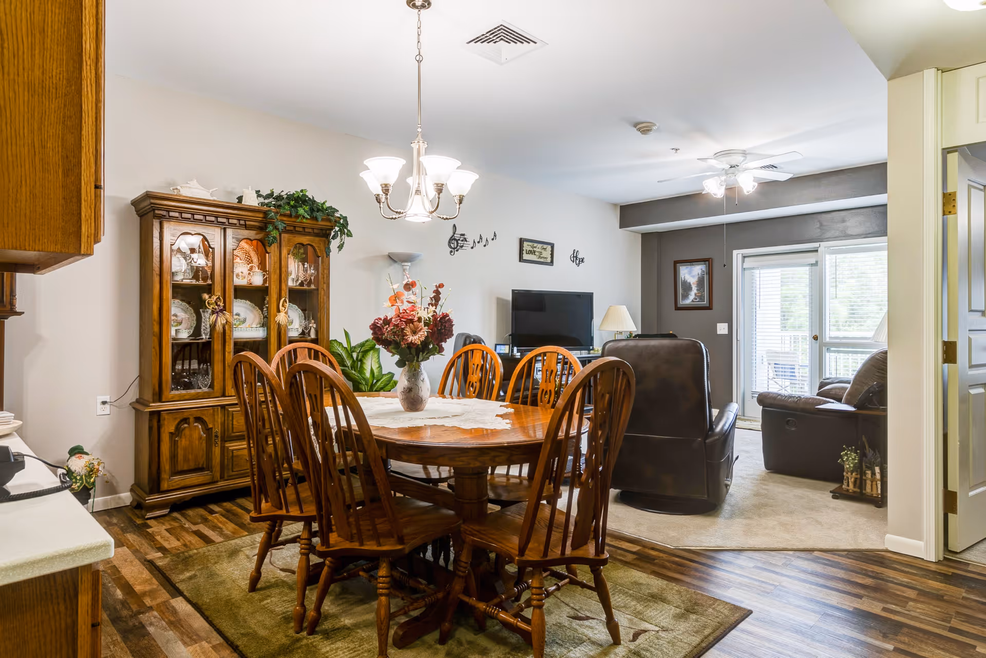 A cozy dining and living area in a senior living facility with a wooden dining table and six chairs, a floral centerpiece on the table, a wooden china cabinet with dishes, and a living room with leather recliners, a TV, ceiling fan, and a sliding glass door leading to a balcony.