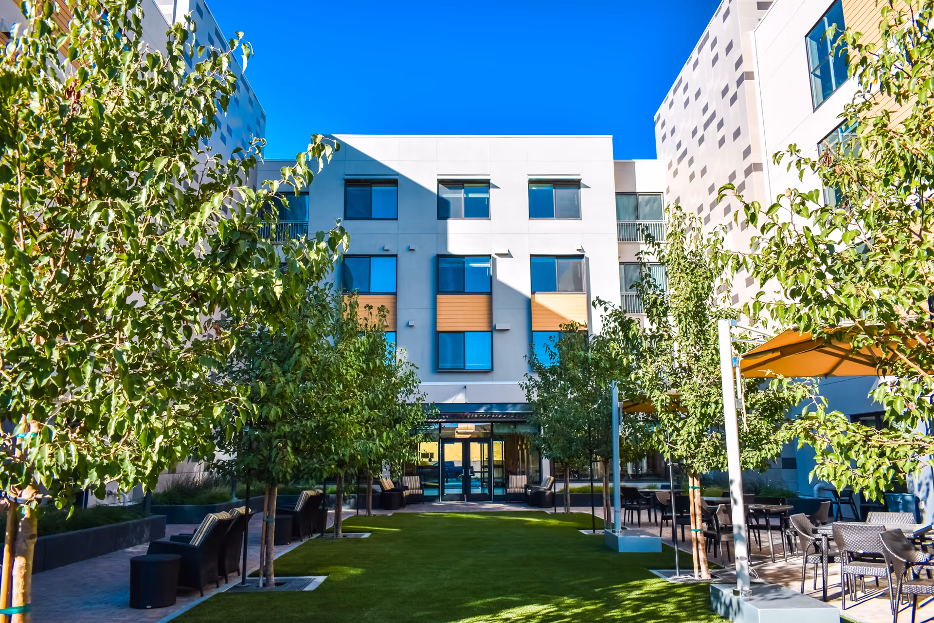 Outdoor courtyard area of a senior living facility with green grass, small trees, seating areas with chairs and tables, and a modern multi-story building in the background under a clear blue sky.