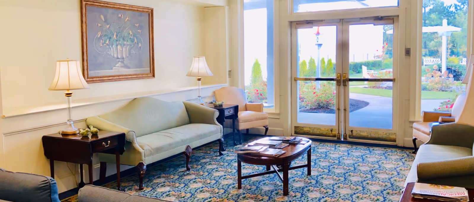 A bright and cozy senior living facility common area with two light green sofas, two beige armchairs, two side tables with lamps, a wooden coffee table with magazines, a floral patterned carpet, a framed painting on the wall, and large glass doors leading to a garden outside.