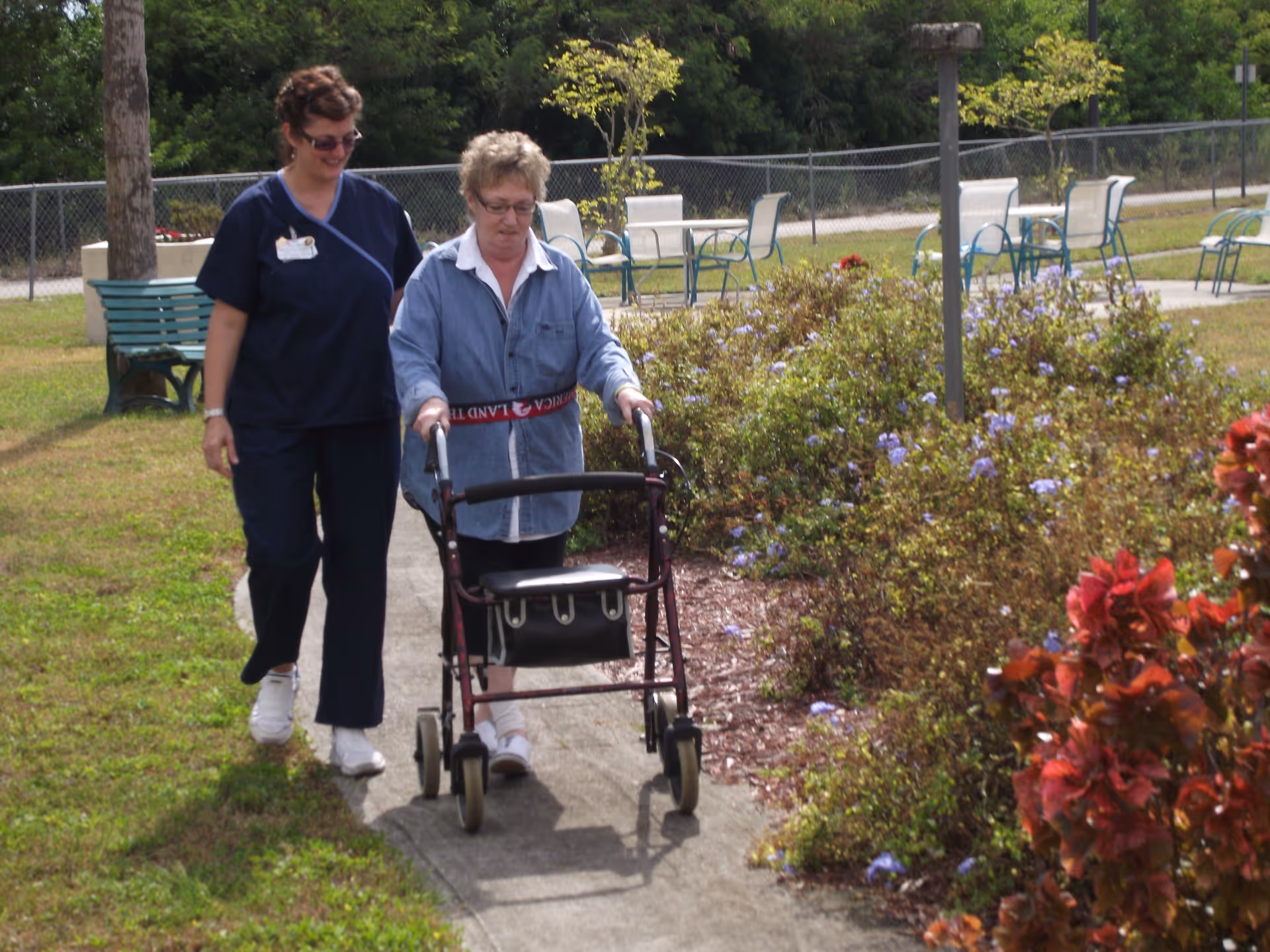 An elderly woman using a walker is being assisted by a caregiver as they walk along a garden path with green grass, flowering bushes, and outdoor chairs and tables in the background.