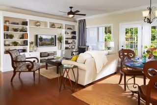 A bright and spacious living room with a white sofa, wooden armchair, coffee table, and built-in shelves filled with decorative items. There is a ceiling fan, large windows with curtains, and a dining table with chairs to the right side near a glass door leading outside.