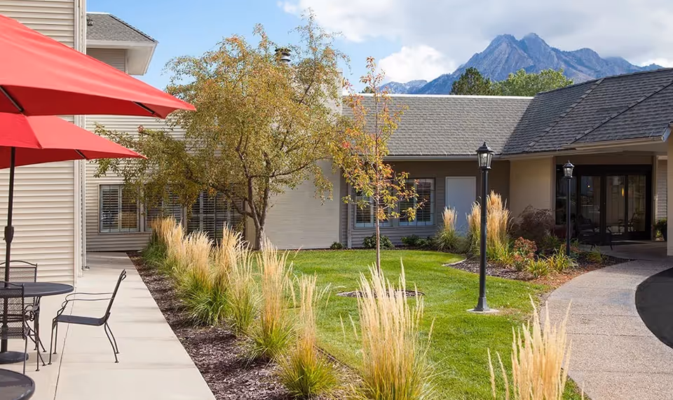 Outdoor courtyard area of a senior living facility with a paved walkway, green lawn, ornamental grasses, trees, and patio tables with red umbrellas. The building surrounds the courtyard and mountains are visible in the background under a partly cloudy sky.