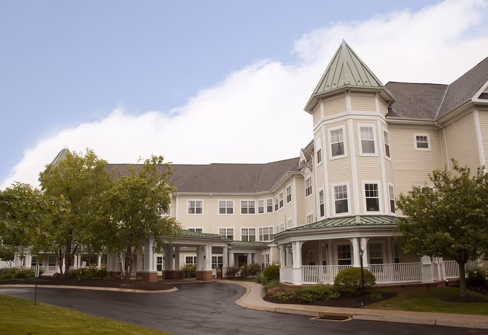 Exterior view of a large, multi-story senior living facility with beige siding, numerous windows, and a green metal roof. The building features a turret-like structure and a covered entrance driveway surrounded by trees and landscaped greenery under a partly cloudy sky.
