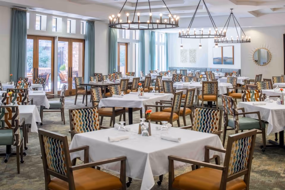 A spacious dining room with multiple tables covered in white tablecloths, each set with glasses, napkins, and small flower vases. The chairs have patterned upholstery with a mix of colors. Large windows and glass doors let in natural light, and modern chandeliers hang from the ceiling. Light blue curtains frame the windows, and there are decorative wall pieces and a round mirror on the walls.