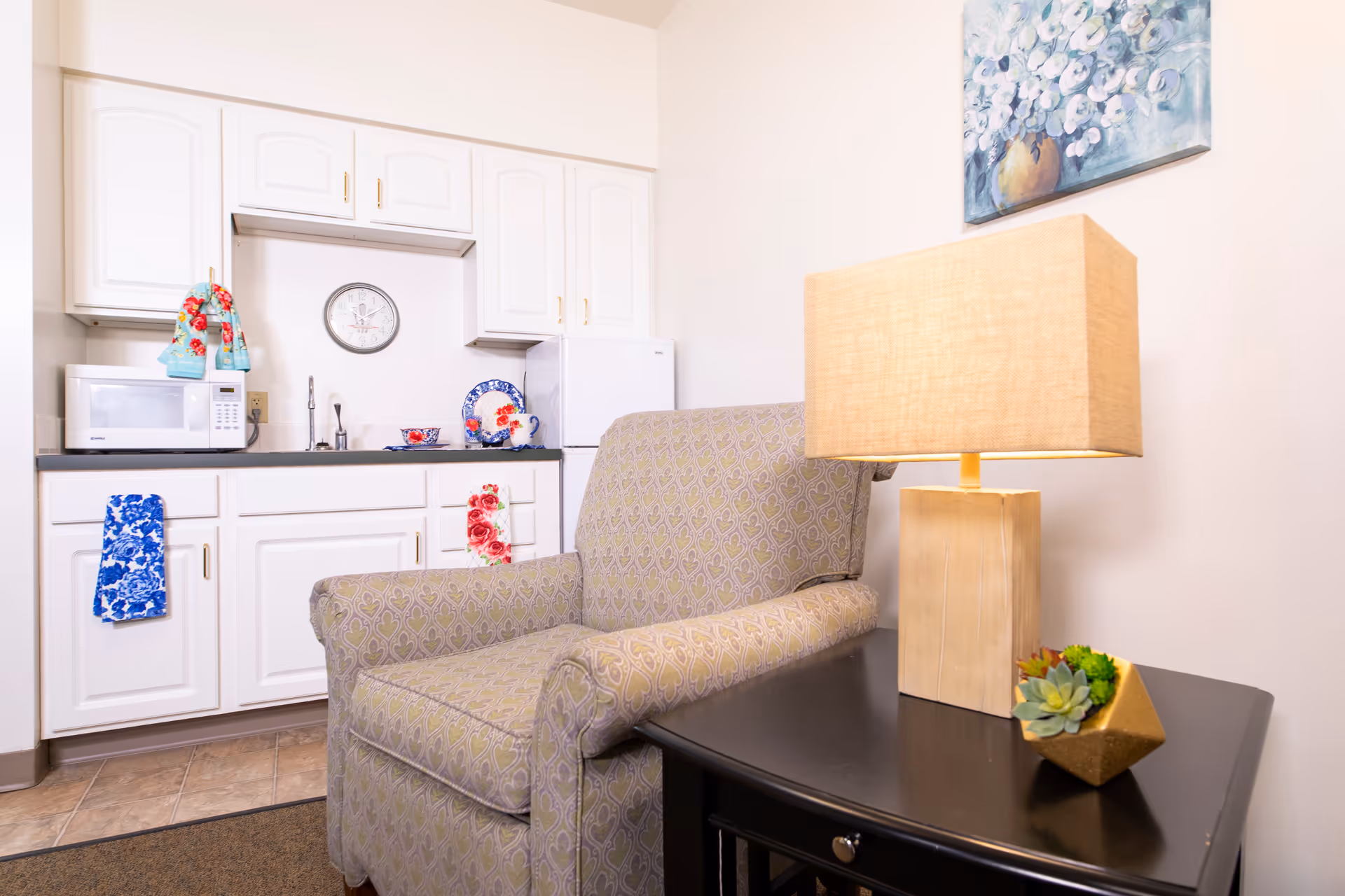 A cozy corner of a room featuring a patterned armchair next to a black side table with a beige rectangular lamp and a small succulent plant. Behind the chair is a kitchenette with white cabinets, a microwave, a small refrigerator, a sink, and decorative towels and dishes. A floral painting hangs on the wall above the lamp.