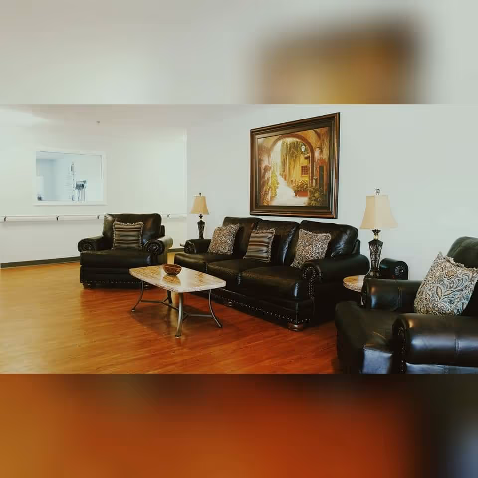 A cozy living room area with dark leather sofa and armchairs, decorated with patterned cushions. A wooden coffee table is placed in front of the sofa. Two table lamps with beige shades are positioned on either side of the sofa. A framed painting depicting an archway and pathway hangs on the white wall behind the sofa. The room has wooden flooring and a large window opening to another room.