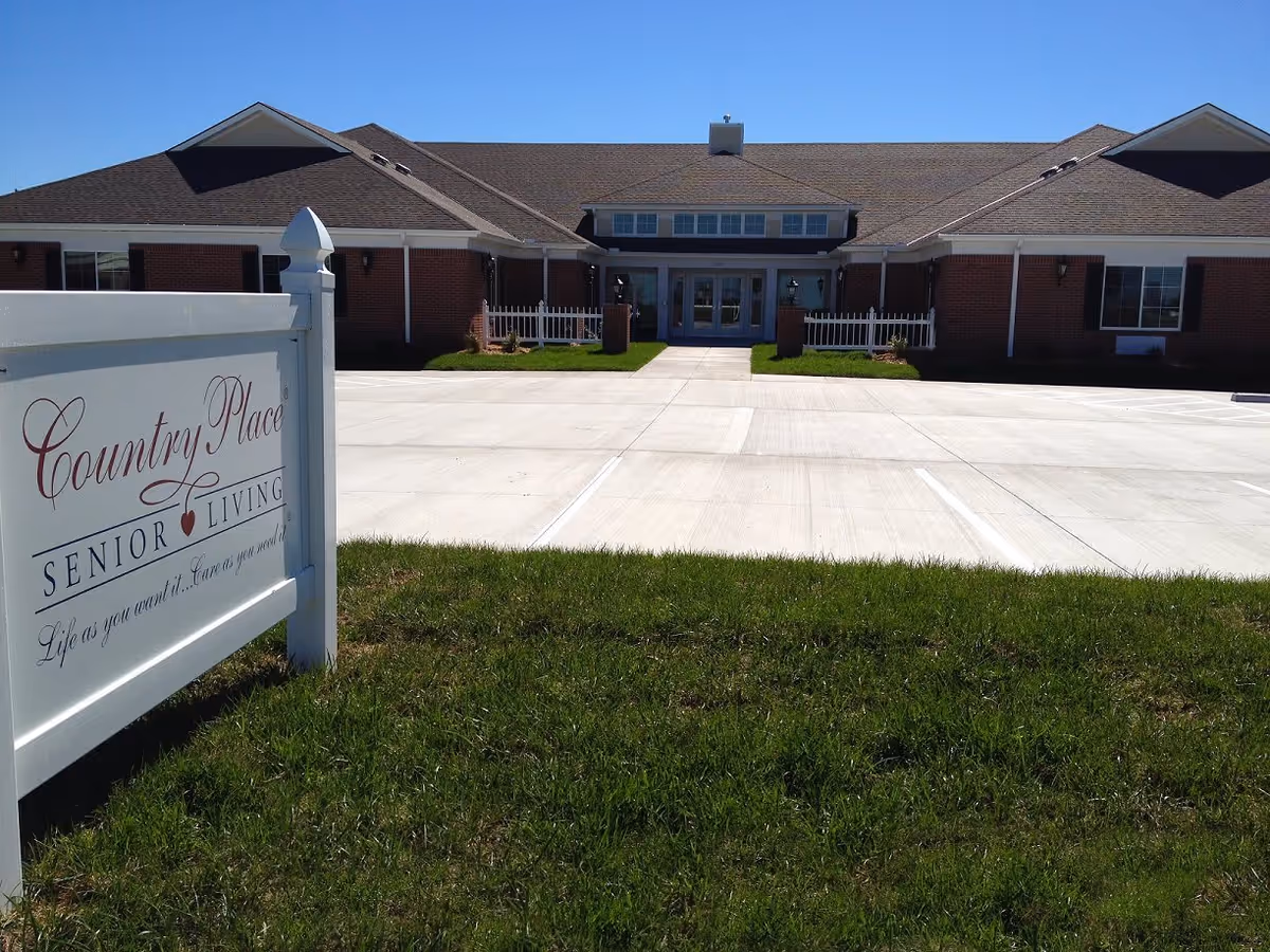 Exterior view of a single-story senior living facility building with a large paved parking area in front and a green lawn. A white sign in the foreground reads 'Country Place Senior Living Life as you want it... Care as you need it.'