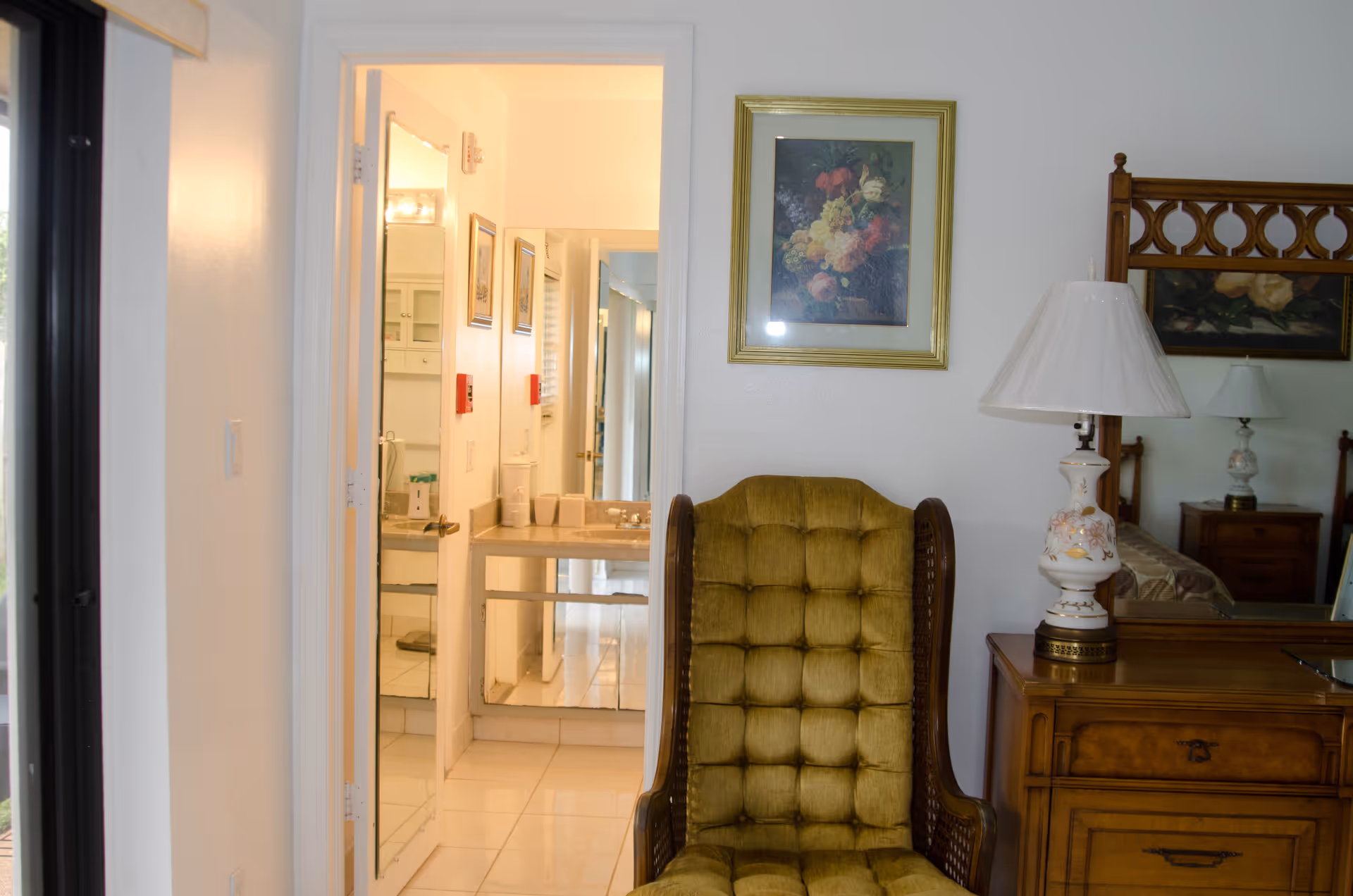 Interior view of a room in a senior living facility showing a green cushioned armchair in front of a wooden dresser with a floral lamp on it. A large mirror is behind the dresser, reflecting part of a bed and another lamp. A framed floral painting hangs on the wall above the chair. To the left, an open door reveals a bathroom with a sink, mirror, and tiled floor.
