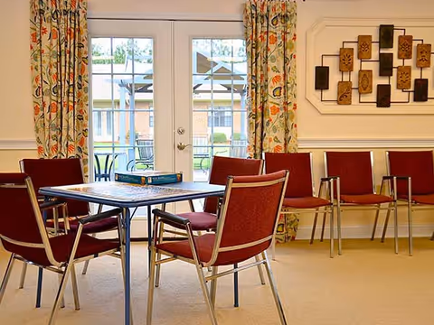 A bright communal room with a square table surrounded by red upholstered chairs facing glass double doors to an outdoor patio.