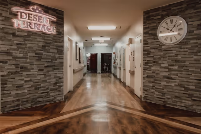 A well-lit hallway in Desert Terrace Healthcare Center with wood-patterned flooring and gray stone-textured walls. On the left wall, there is a lit sign that reads 'DESERT TERRACE.' A large white clock with Roman numerals is mounted on the right wall. Several doors and framed pictures line the hallway, and a walker is visible at the far end near an exit door.