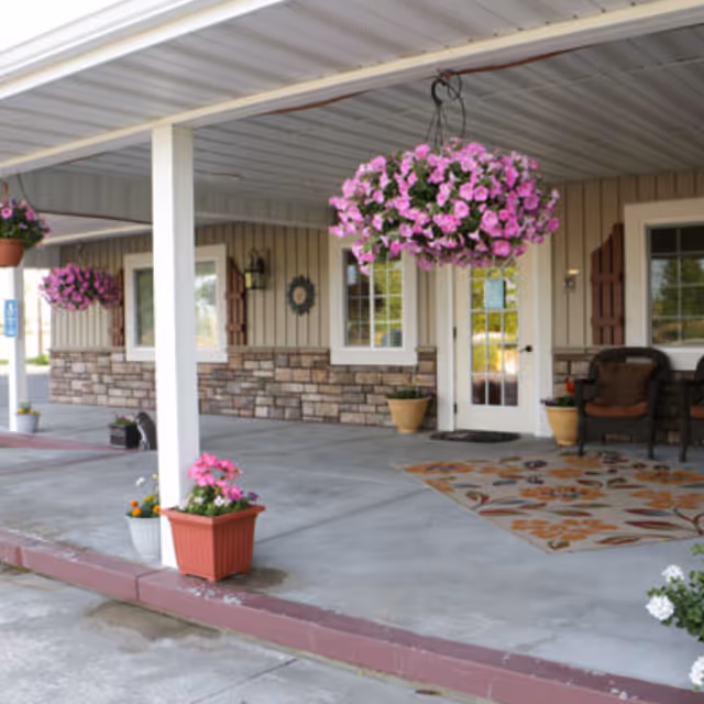 Covered outdoor patio area with hanging baskets of pink flowers, potted plants, a patterned outdoor rug, and seating near the entrance door of a building with stone and wood siding.
