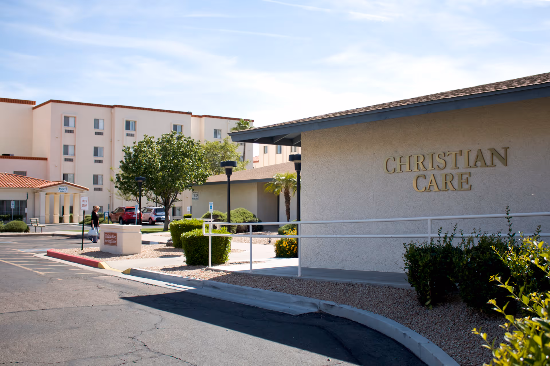 Exterior view of Christian Care Health Center showing a single-story building with the name 'CHRISTIAN CARE' on the wall, a parking lot, landscaped bushes, trees, and a multi-story building in the background under a clear sky.