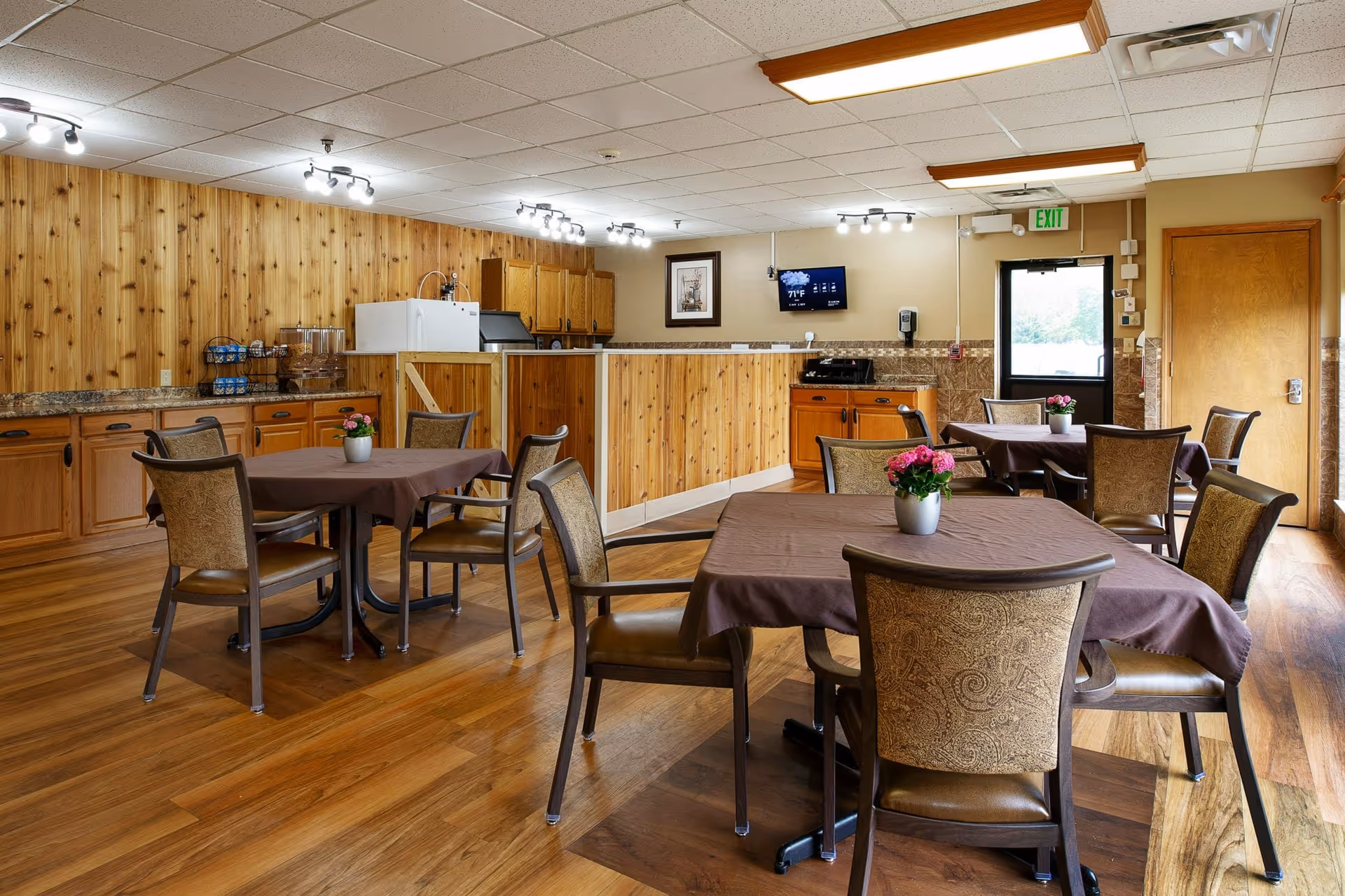 Dining area with several tables and chairs, table centerpieces, and a wood-paneled serving counter and kitchenette.