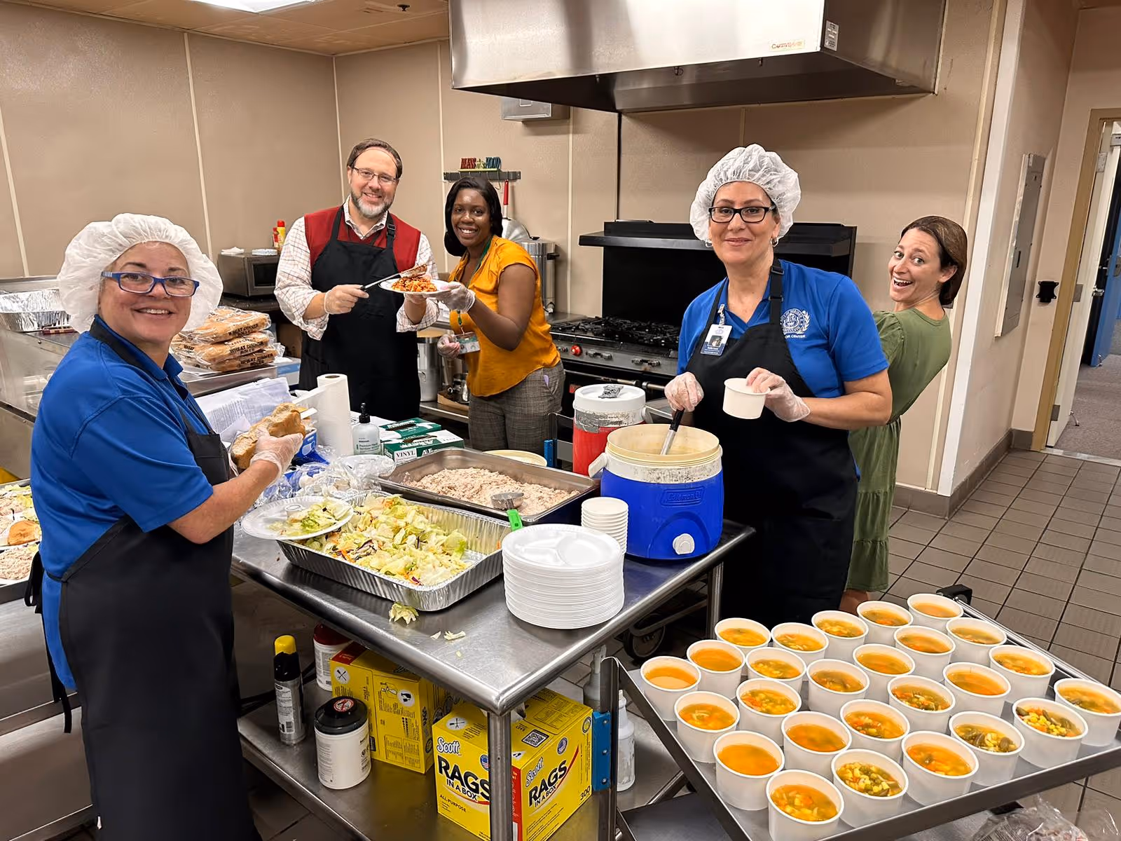 Five people in a kitchen preparing and serving food. Two women in blue shirts and hairnets are serving soup and holding bread, a man in a red vest and apron is serving spaghetti to a woman in a yellow top, and another woman in a green dress is smiling in the background. The kitchen has trays of salad, soup cups, and other food items on a stainless steel table.